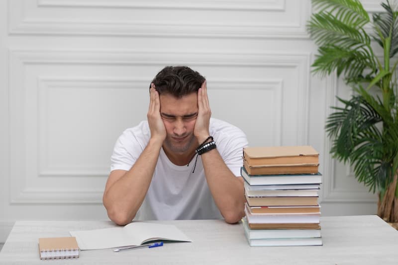 Ein junger Mann sitzt mit verzerrtem Gesicht vor seinen Büchern, wie lässt sich Prüfungsangst überwinden?