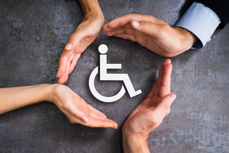 Ein Symbol für einen Menschen mit Behinderung wird von Händen umkreist, was bedeutet Barrierefreiheit im Job?